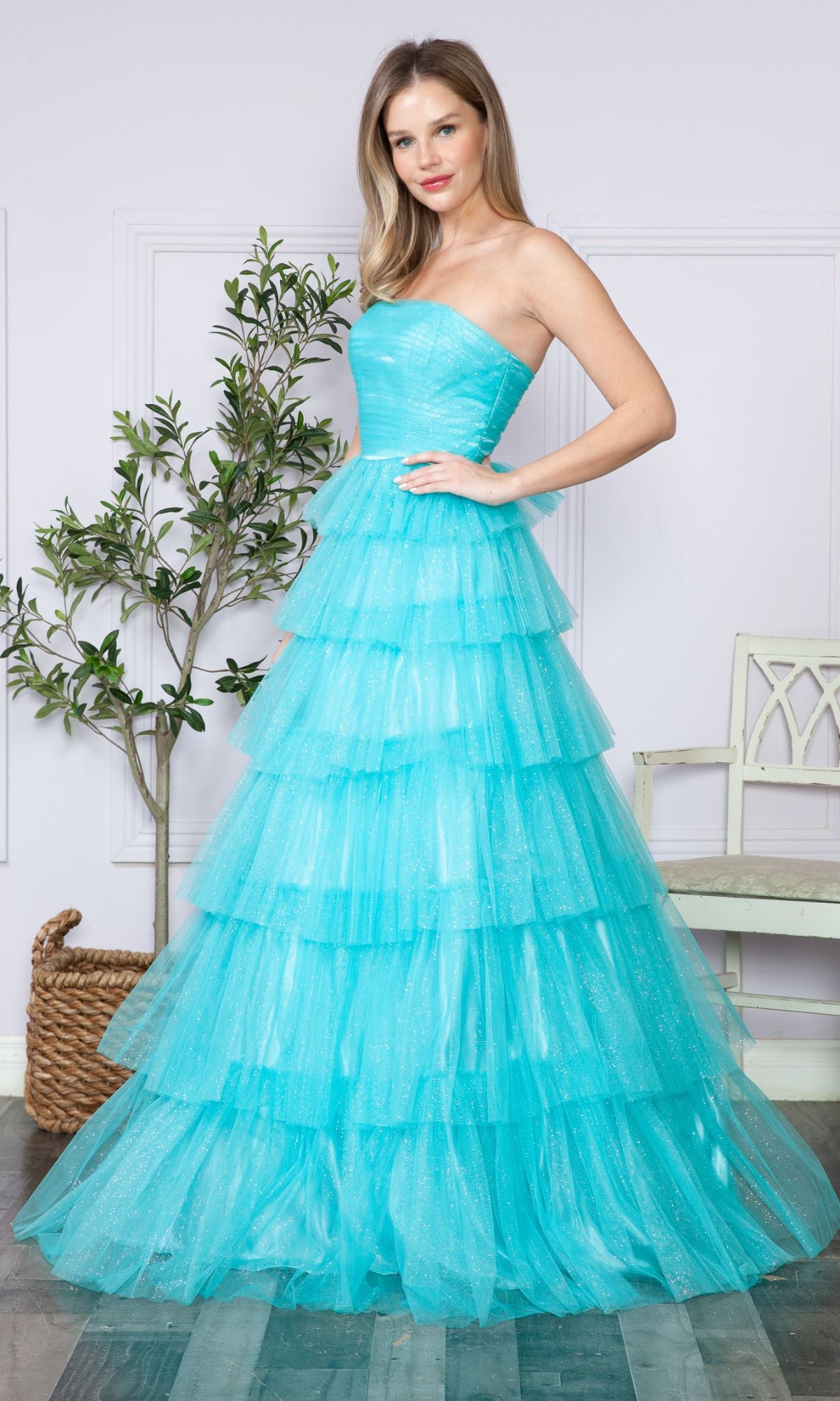A woman stands indoors wearing the Strapless Long Tiered Glitter Prom Ball Gown 9386 in turquoise tulle; tiered ruffles cascade down, with a potted plant and white chair in the background.